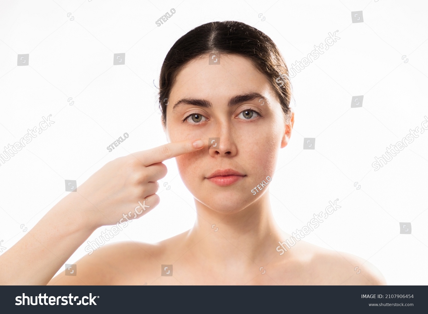 Plastic nose surgery. Portrait of young caucasian woman point her crooked bridge of the nose isolated on white background. Copy space. Concept of rhinoplasty. #2107906454