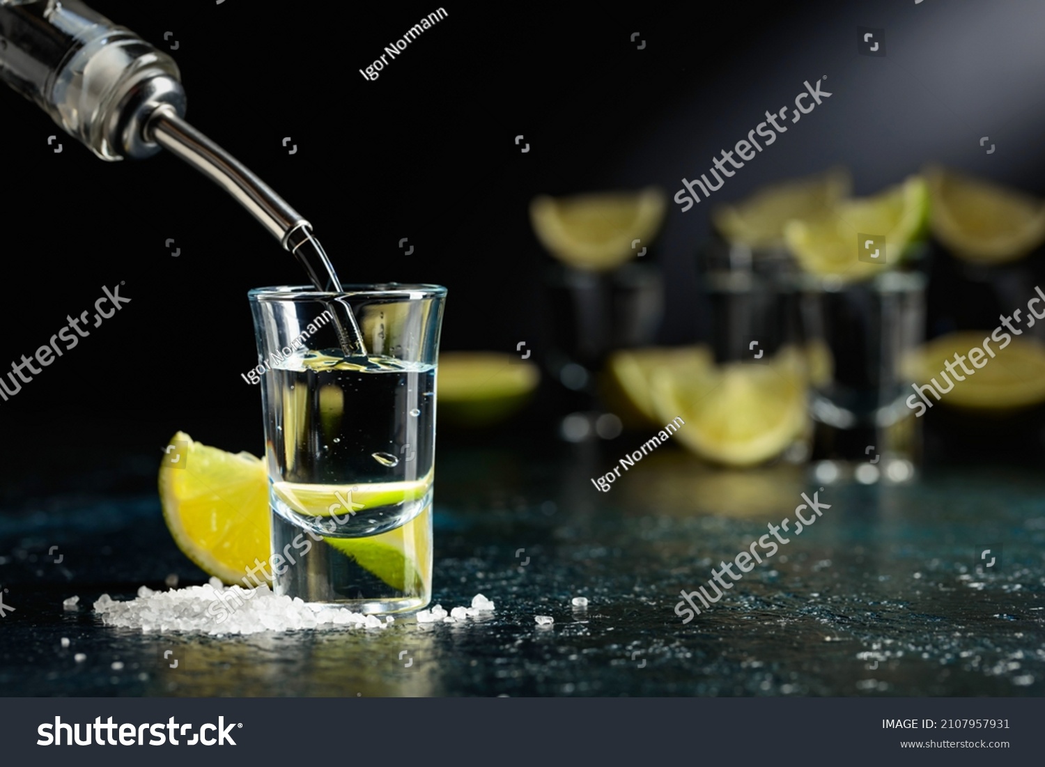 Tequila is poured into a glass. Tequila shots with lime slices and sea salt on a dark blue table. #2107957931