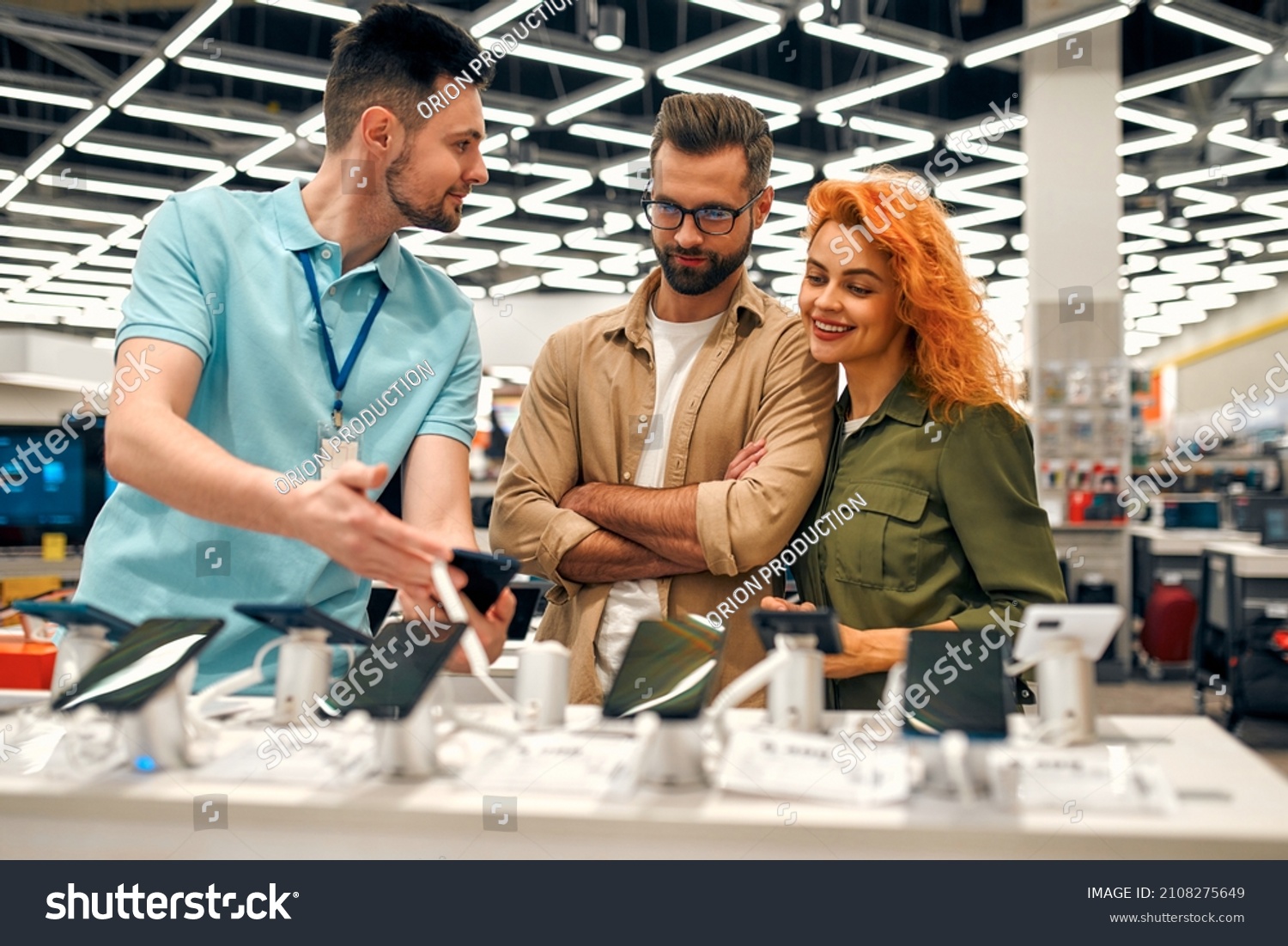 Red-haired sweet woman, along with her boyfriend, chooses new smartphone in store of household appliances, electronics and gadgets, getting help from store consultant. #2108275649