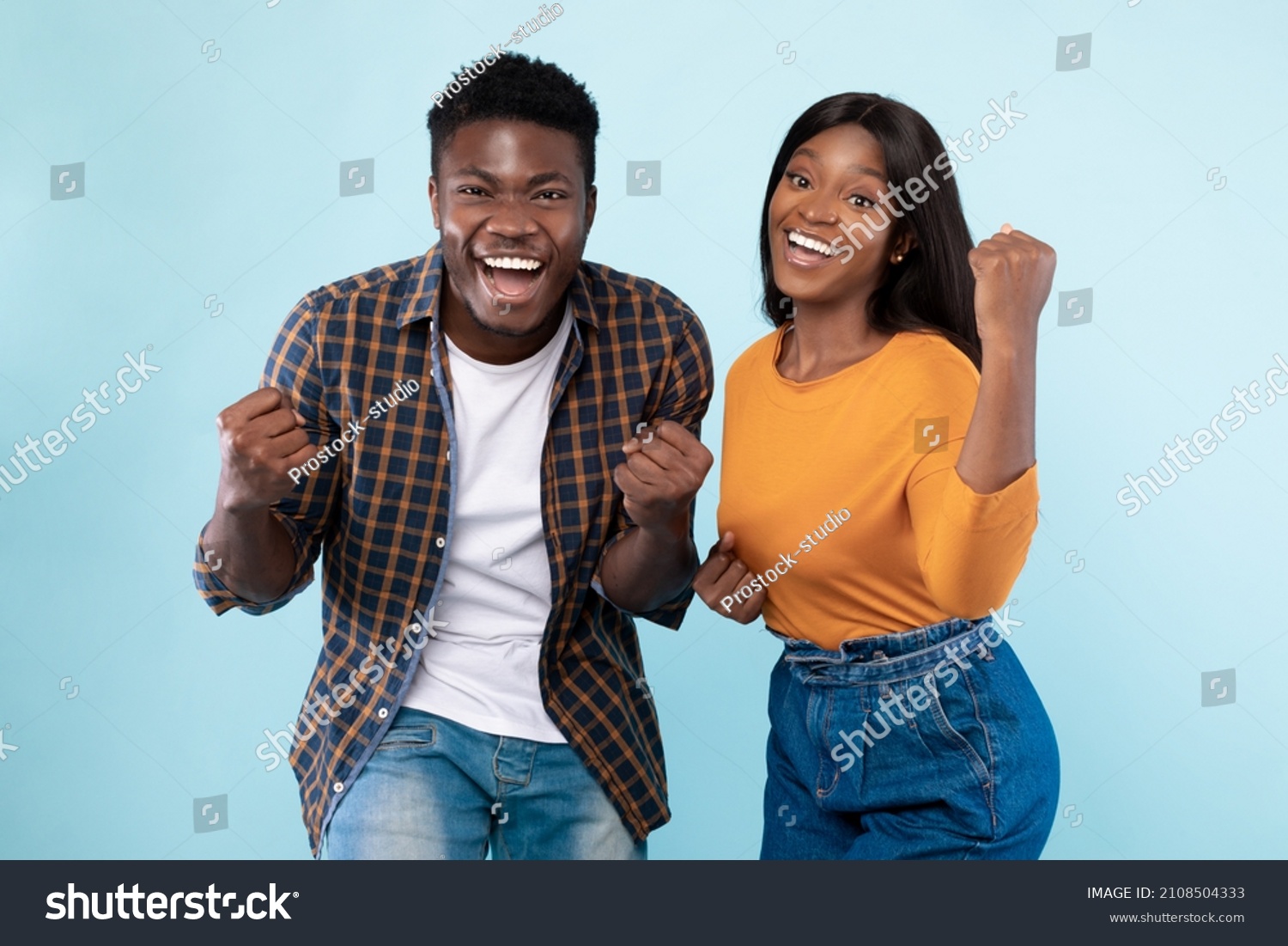 Wow, Yes. Portrait of overjoyed young black man and woman cheering and shaking clenched fists, looking at camera. Happy couple celebrating win posing standing isolated over blue studio background wall #2108504333