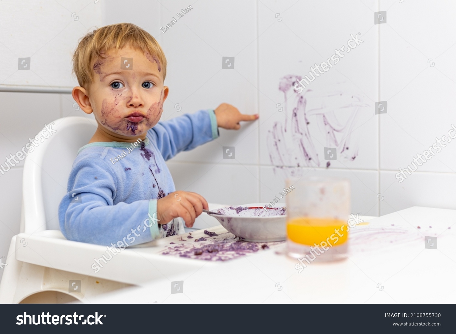 blond boy in his high chair shows the wall that he has stained with his breakfast #2108755730