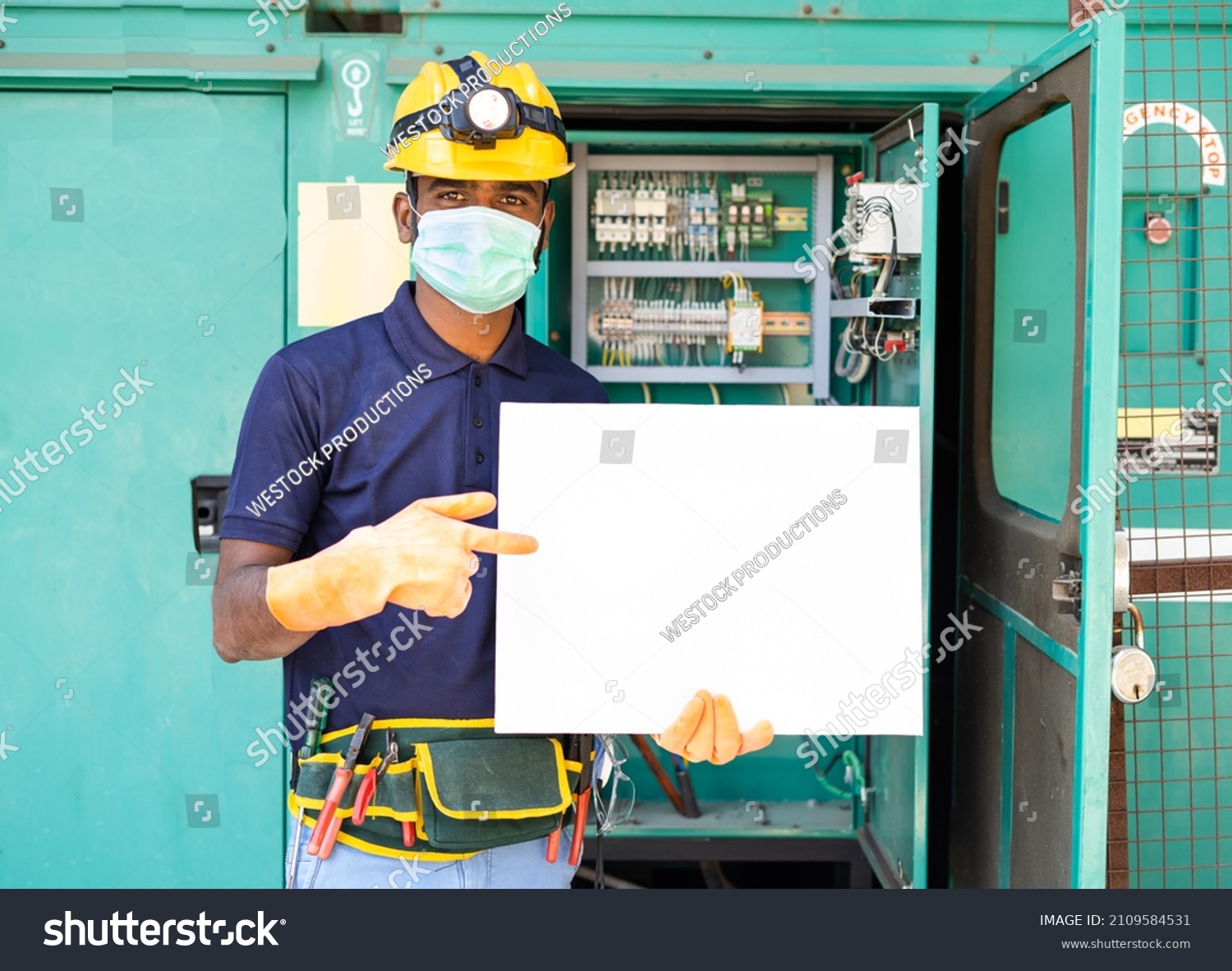 Electrician with meidcal face mask and safety electrician with meidcal face mask and safety precautions holding empty sign board by looking at camera - concept of advertisement,promotion and job offer #2109584531