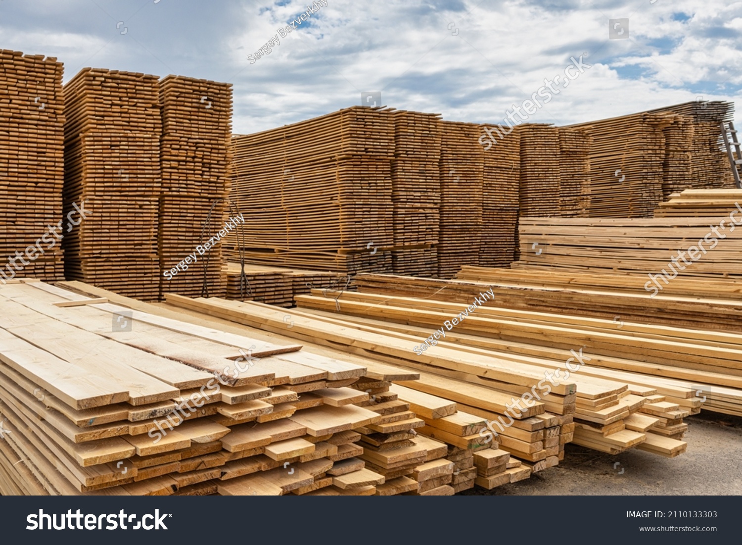 Industrial production of finished lumber products stacked in stacks on the warehouse territory. On an open background of blue sky with clouds. #2110133303