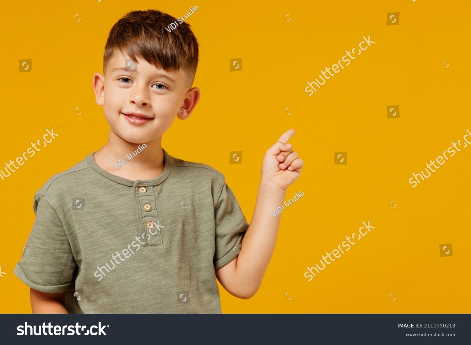Little small happy boy 6-7 years old in green casual t-shirt point index finger aside on workspace area isolated on plain yellow background studio portrait. Mother's Day love family lifestyle concept. #2110550213