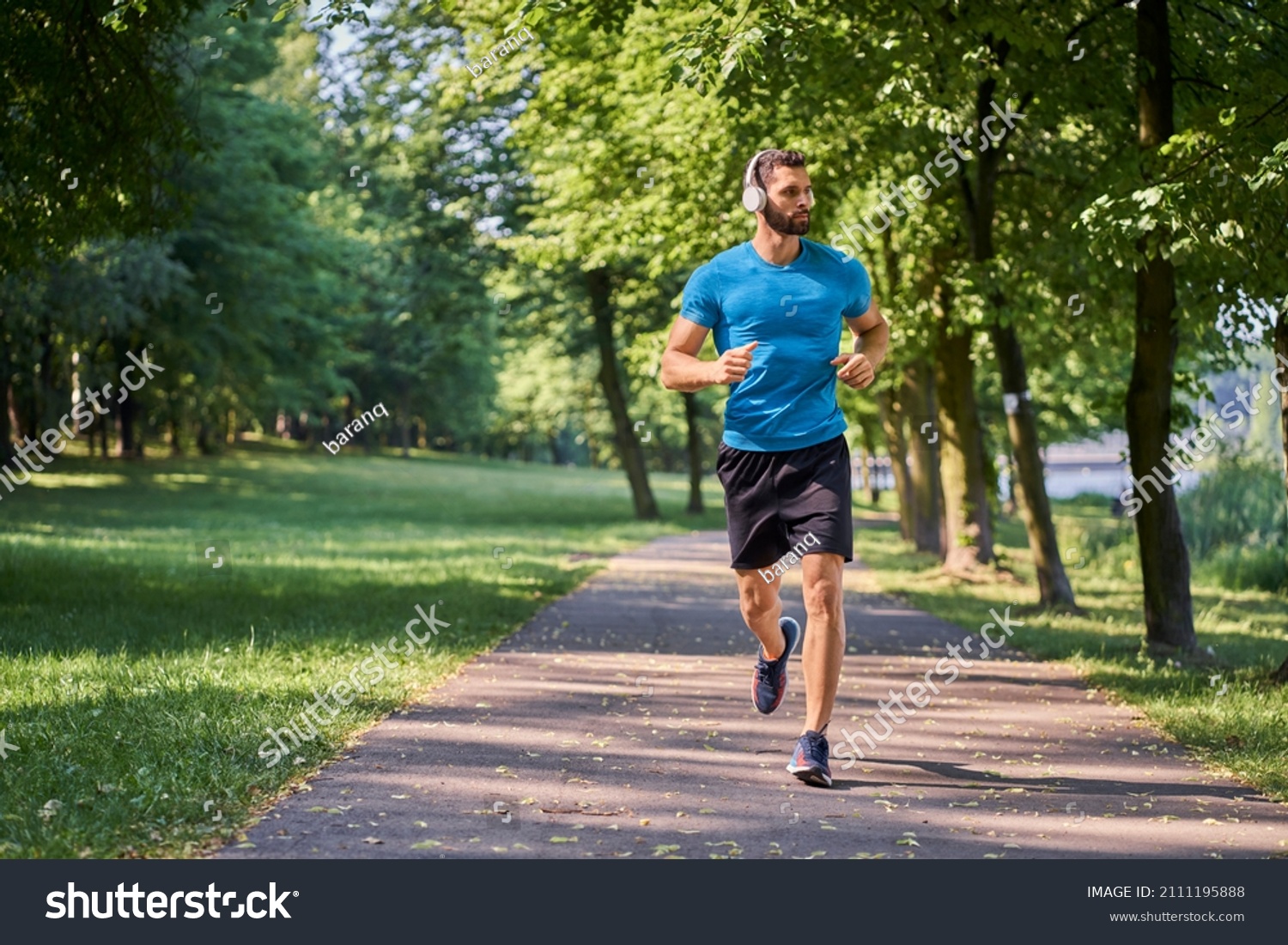 Young man running in park with headphones on sunny summer day #2111195888