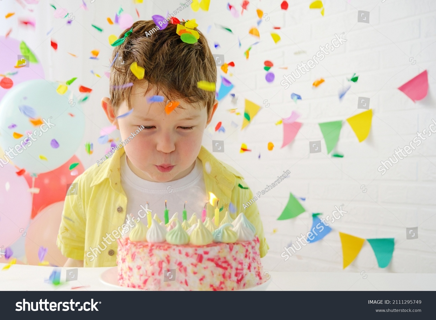 The child blows out the candles on the birthday cake. Boy 5 years old in a festive setting. Birthday, confetti fall on the boy. Horizontal photo. #2111295749