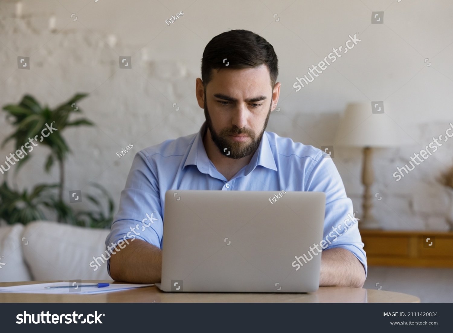 Focused millennial business man using laptop at table. Young entrepreneur, freelancer in casual, employee, worker using compute at home office workplace, typing, chatting online #2111420834