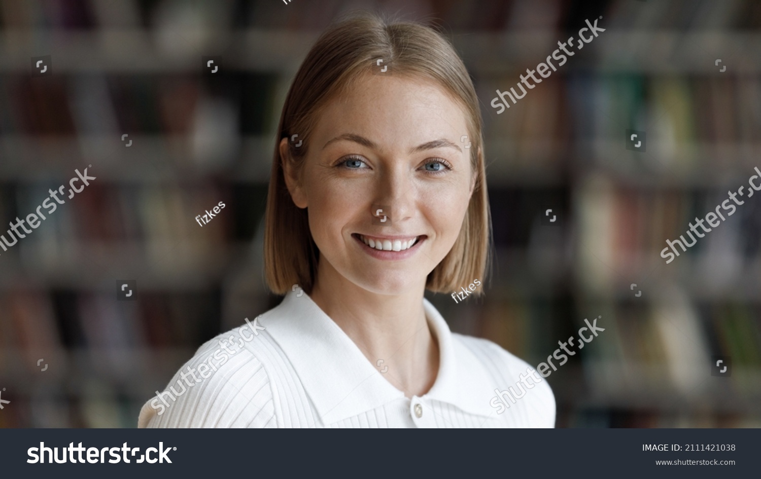 Happy pretty college student girl posing in university library with blurred bookshelves behind. Young millennial woman looking at camera with toothy smile head shot portrait #2111421038