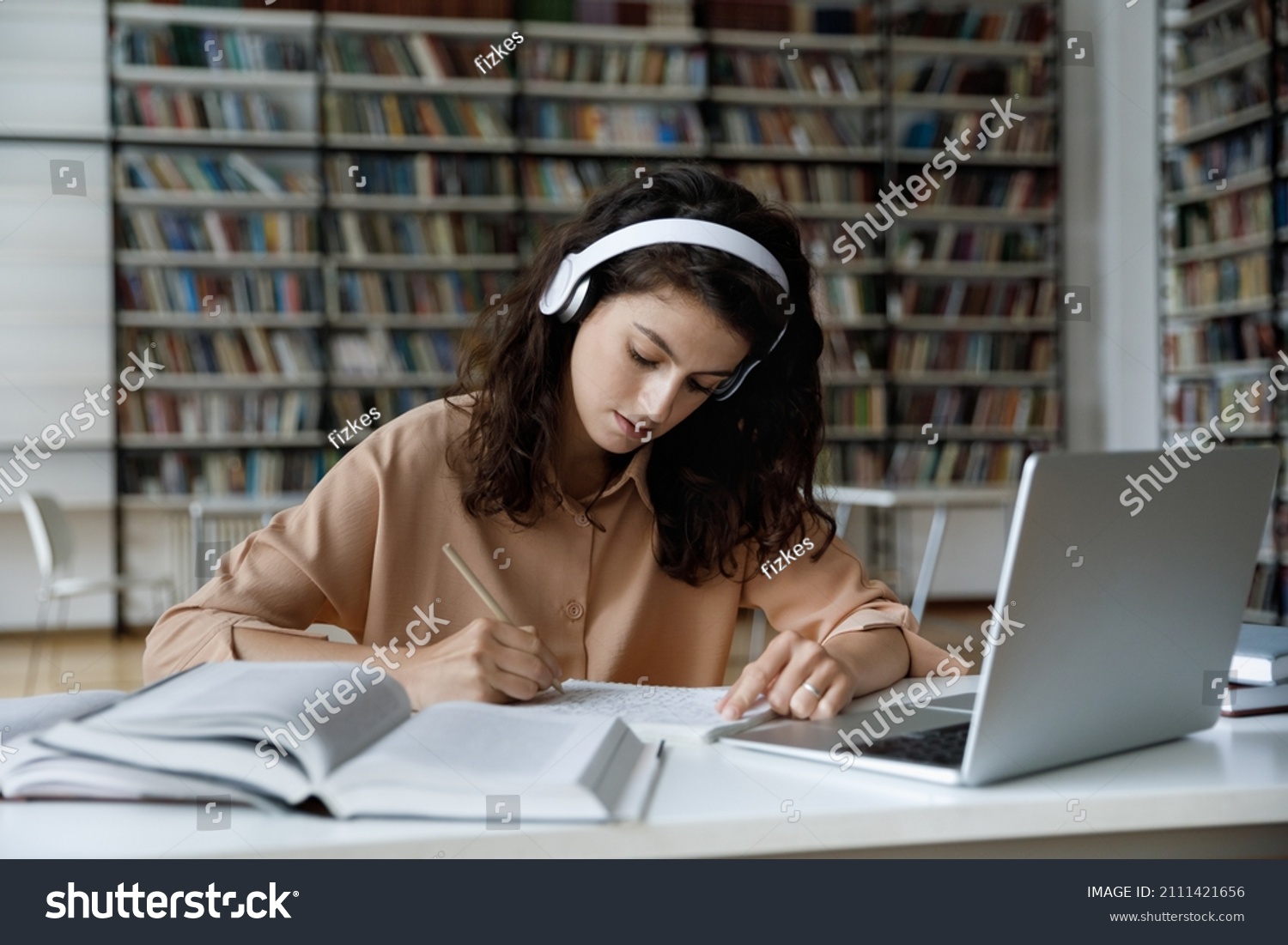 Serious busy hardworking student girl in headphones working essay, study project in college public library, attending online learning conference, watching video lesson on internet, writing notes #2111421656