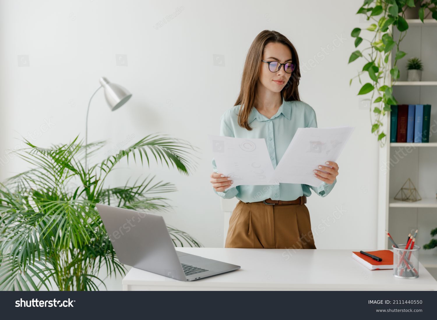 Young successful employee business woman in blue shirt hold paper account documents work stand at workplace white desk with laptop pc computer at light modern office indoors Achievement career concept #2111440550