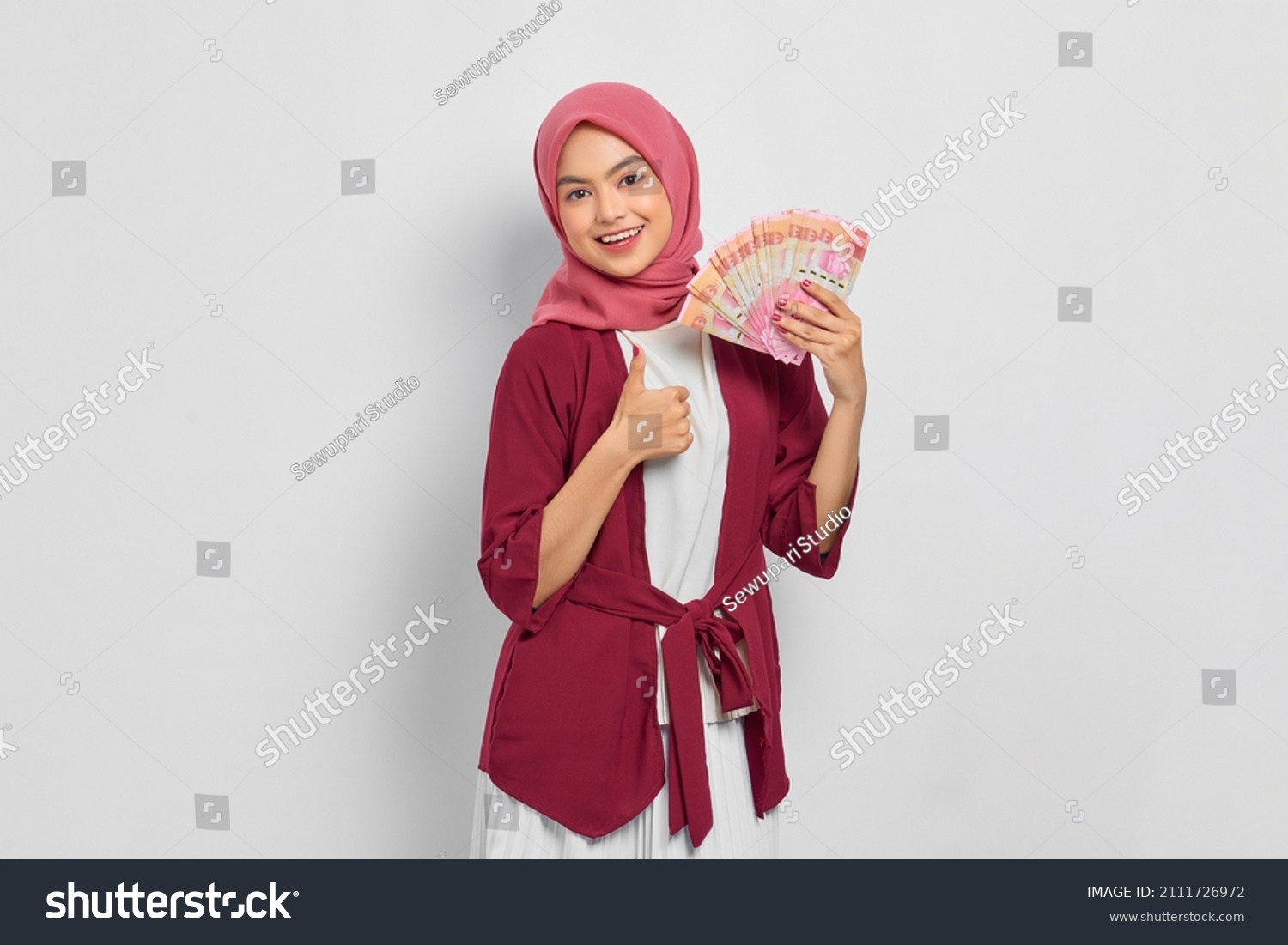 Cheerful beautiful Asian woman in casual shirt and hijab holding Indonesian rupiah banknotes, showing thumb up gesture isolated over white background. People religious lifestyle concept #2111726972