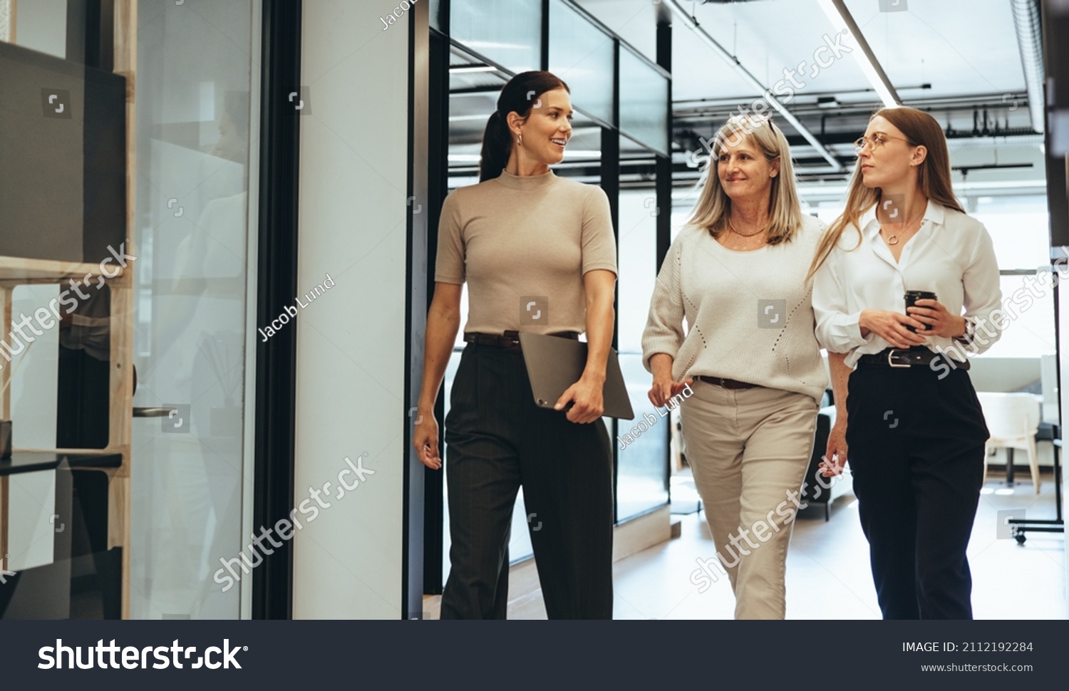 Three cheerful businesswomen walking together in an office. Diverse group of businesswomen smiling while having a discussion. Successful female colleagues collaborating on a new project. #2112192284