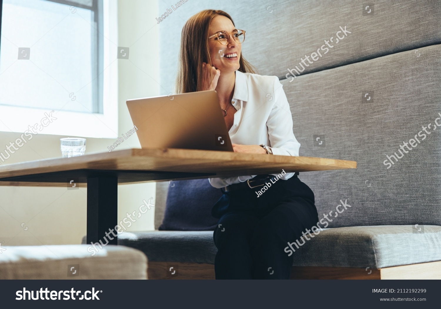 Excited businesswoman looking away thoughtfully. Happy young businesswoman smiling while contemplating business ideas. Young female entrepreneur working on a laptop in a modern workspace. #2112192299
