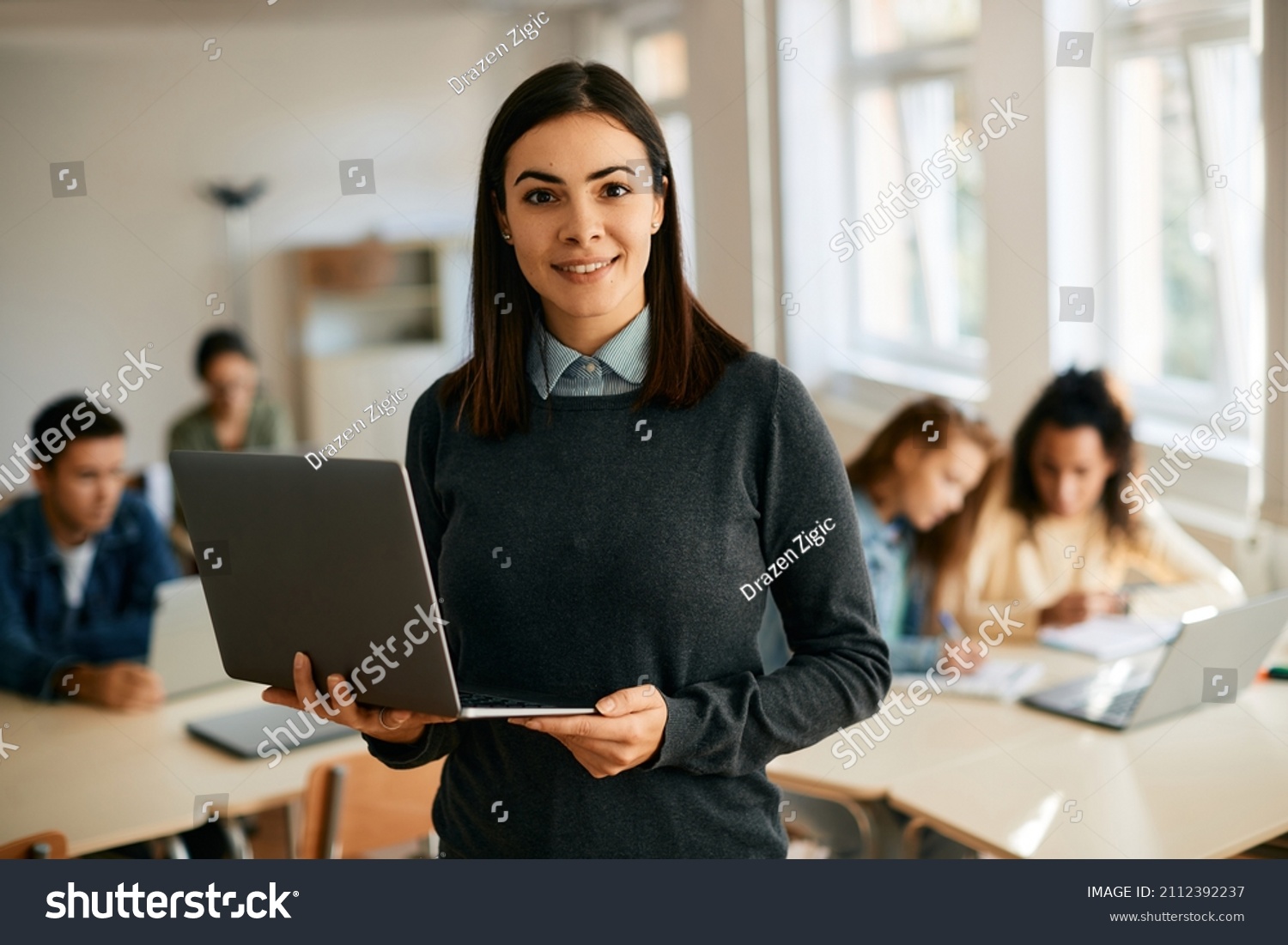 Smiling teacher using laptop during computer class at high school and looking at camera. Her students are learning in the background.  #2112392237