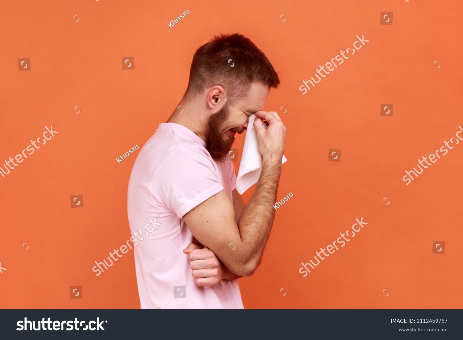 Portrait of sad alone hopeless bearded man standing, holding his head down and crying, wipes tears with handkerchief, wearing pink T-shirt. Indoor studio shot isolated on orange background. #2112459767