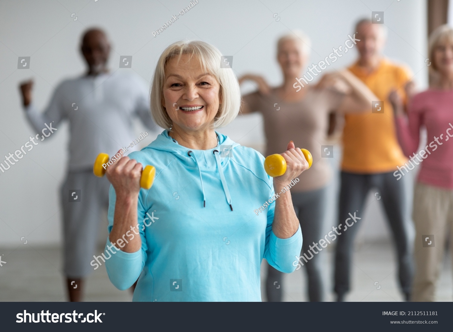Multiracial group of senior people in sportswear doing strength building fitness exercises with dumbbells, holding fitness tools and smiling at camera, selective focus on positive elderly lady #2112511181