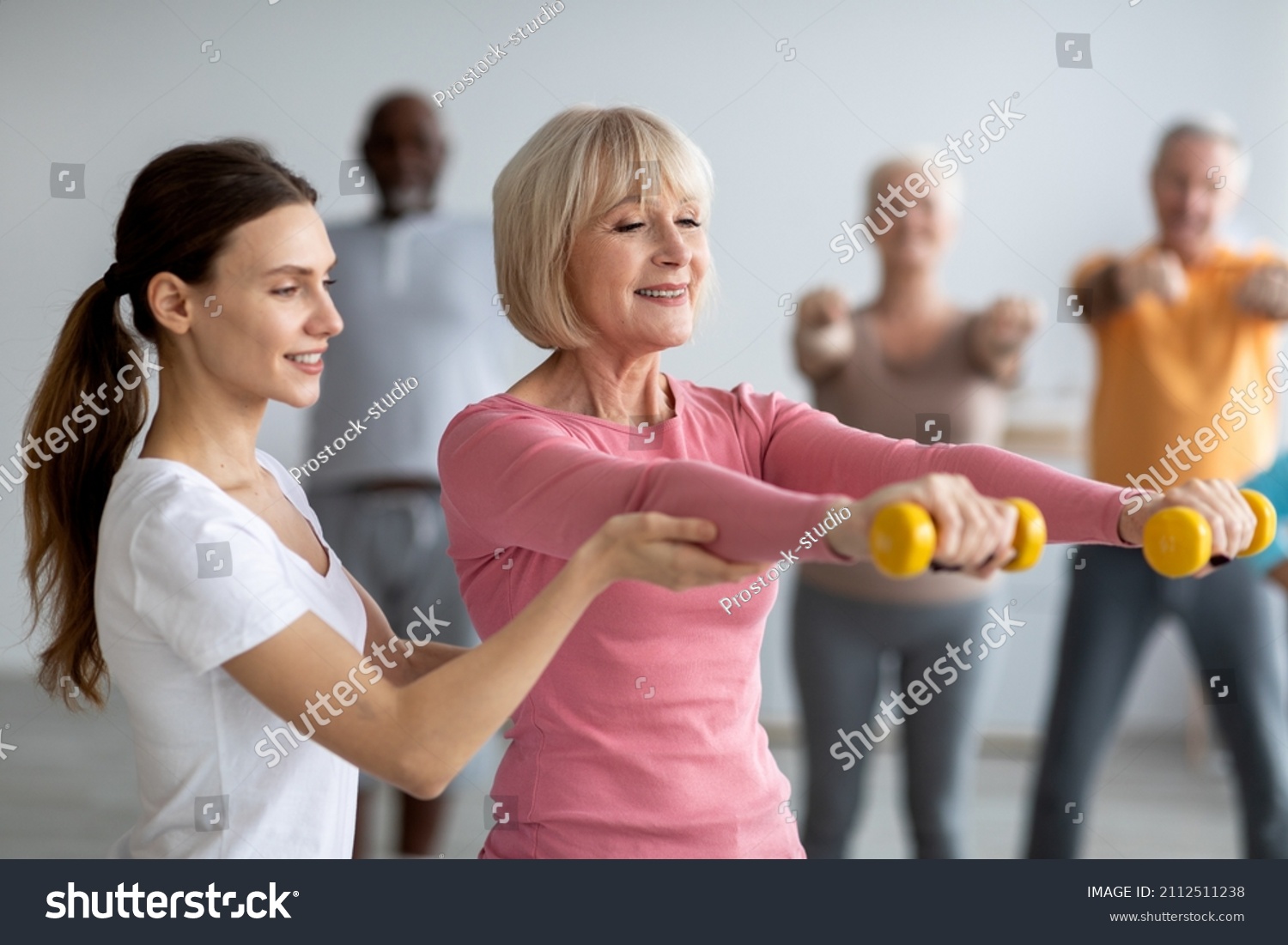 Young lady coach helping senior woman while exercising with dumbbells, sporty multiracial group of elderly people having fitness class at nursing home, training with instructor #2112511238
