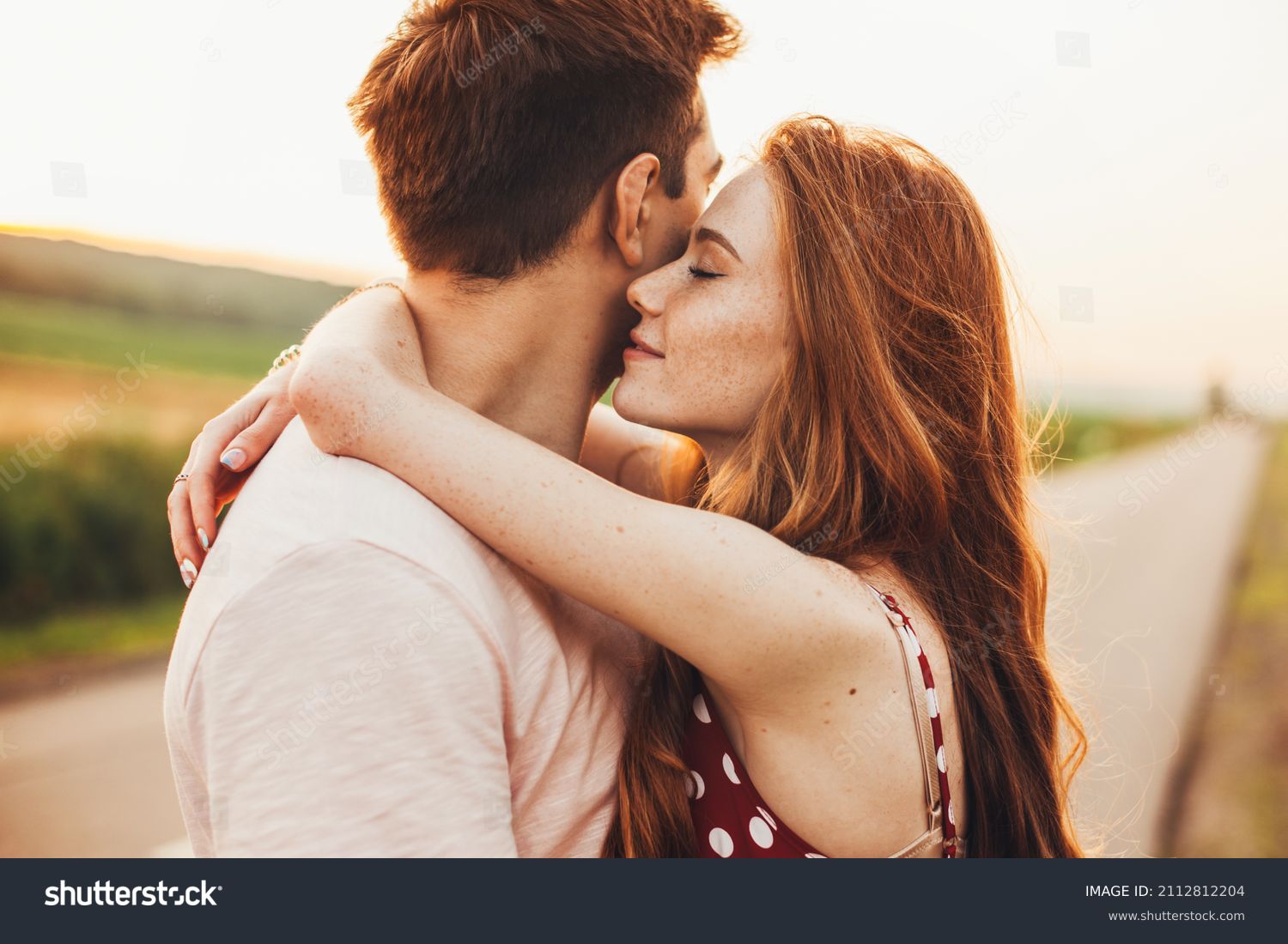 Close-up portrait of a caucasian young loving couple embracing while standing on a roadside. Couple embracing road travel. Sunset scene. #2112812204