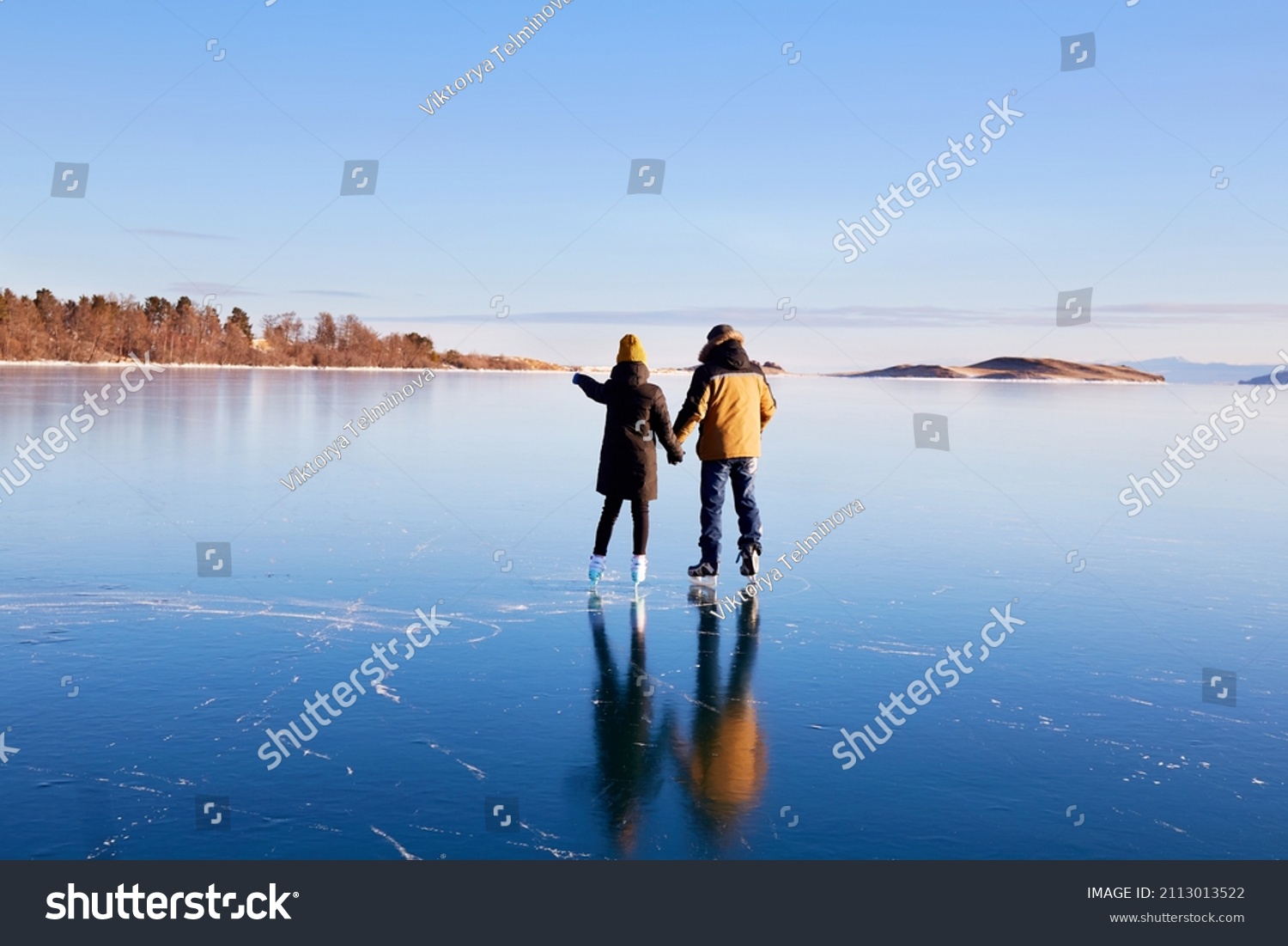 A young couple is skating on the frozen Lake Baikal. Transparent ice. Travel in winter, active recreation, sports.  #2113013522