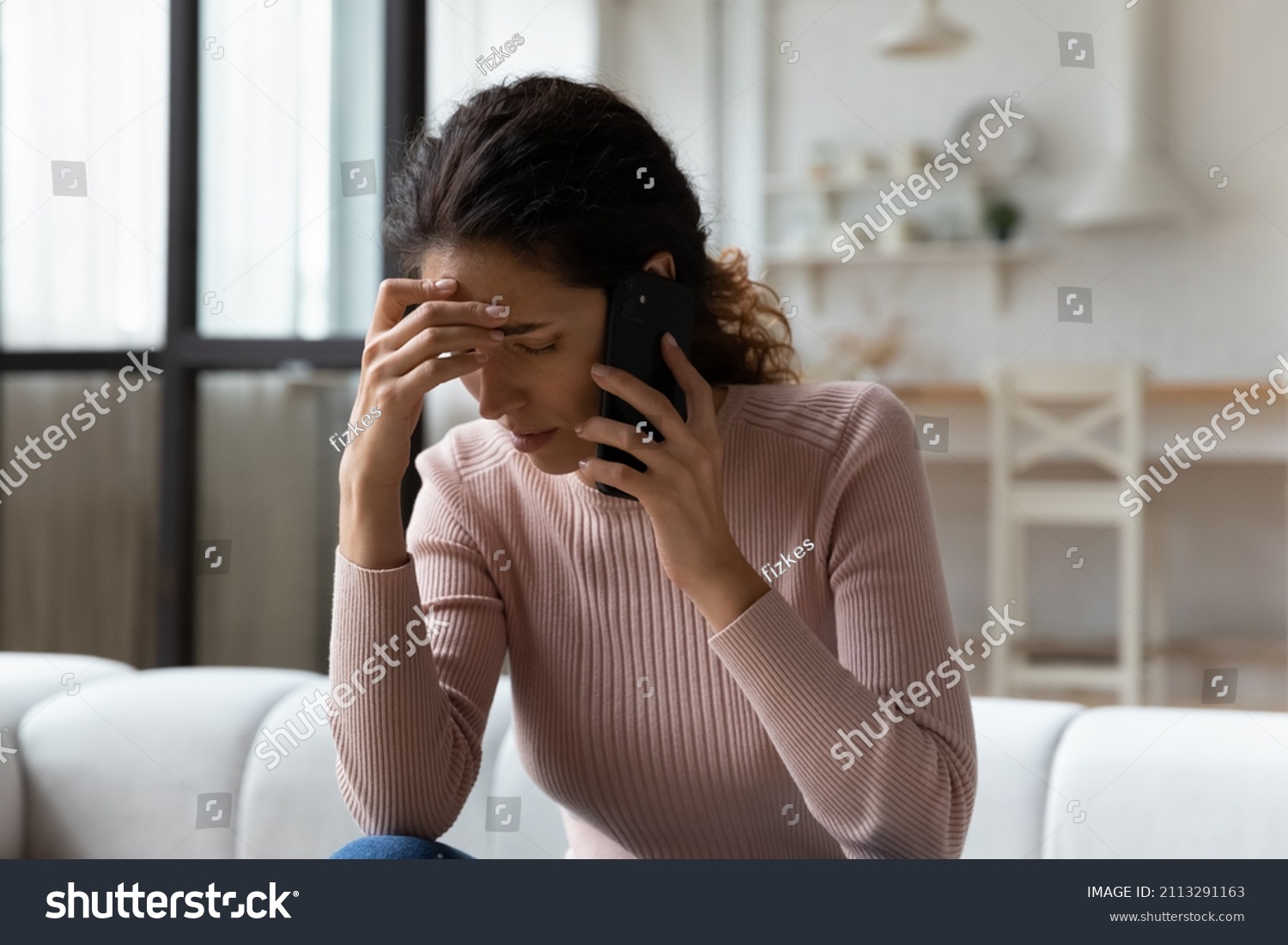 Young Hispanic stressed woman sit on sofa in living room talks on smartphone looking concerned, listen bad news feels desperate, having unpleasant remote conversation, receive disagreeable information #2113291163