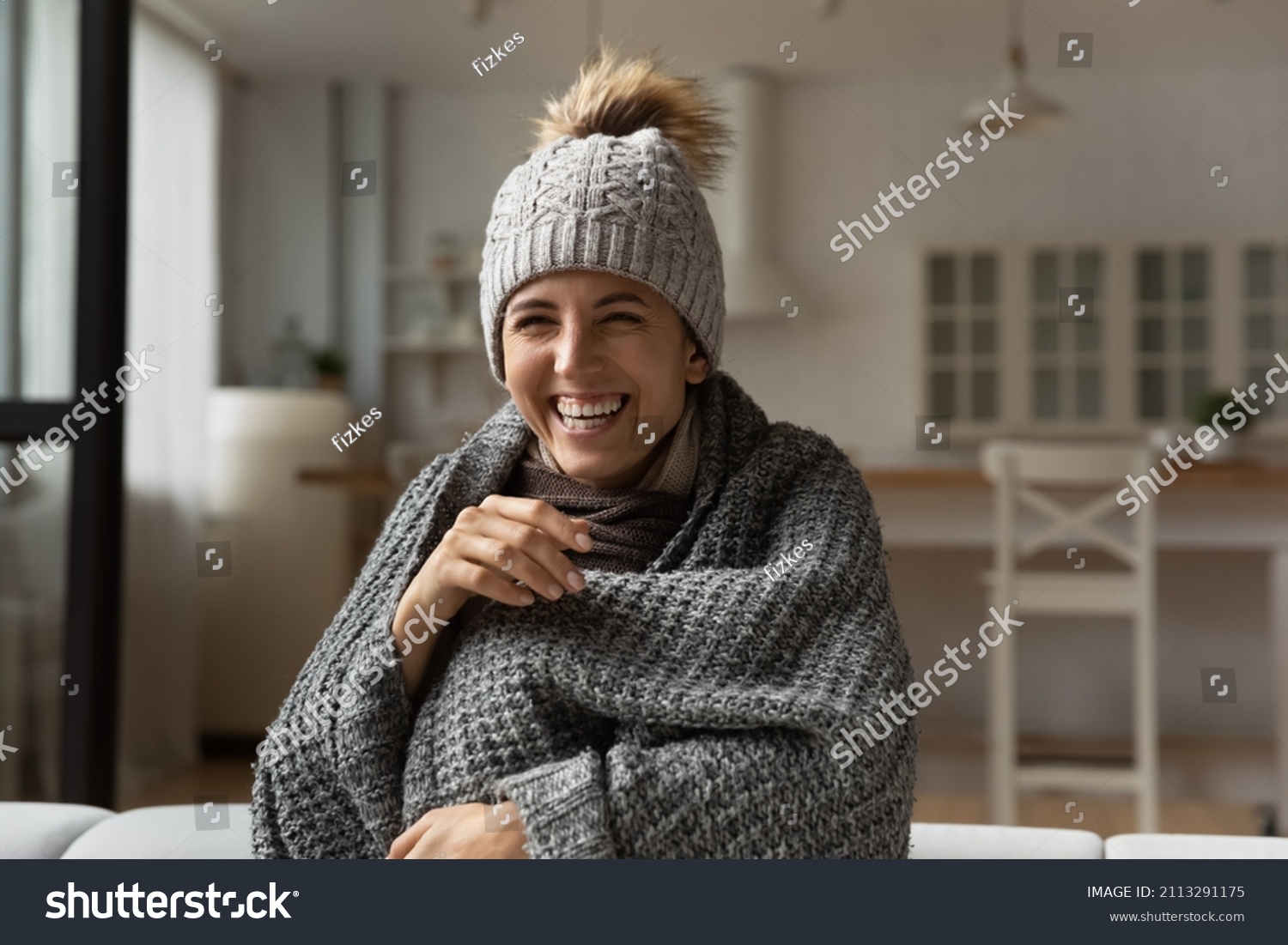 Funny laughing Hispanic woman wear knitted grey bobble hat wrapped in warm plaid sit on sofa relaxing warmup while watch comedy TV show, spend carefree weekend alone at home having fun feel overjoyed #2113291175
