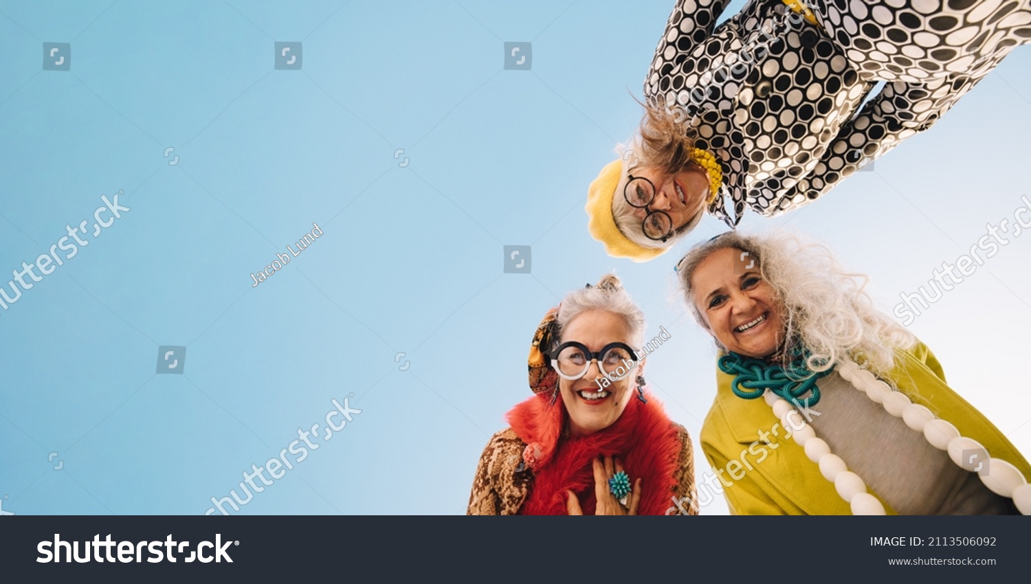 Low angle view of happy senior women smiling at the camera outdoors. Group of cheerful elderly women wearing colourful casual clothing. Three stylish senior women enjoying their golden years. #2113506092