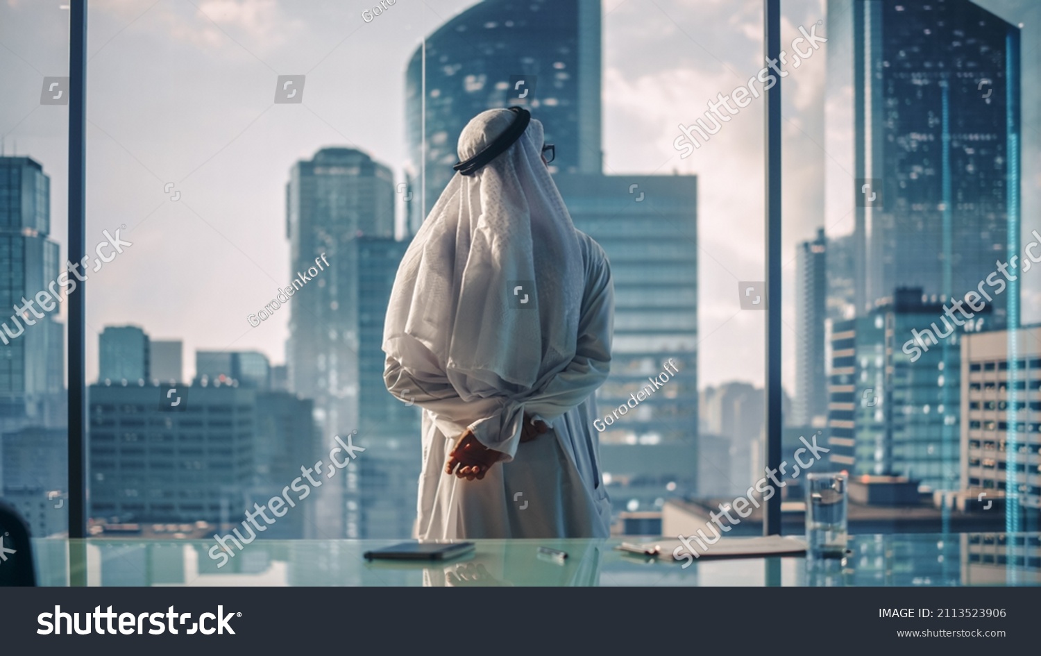 Successful Muslim Businessman in Traditional White Kandura Standing in His Modern Office Looking out of the Window on Big City with Skyscrapers. Successful Saudi, Emirati, Arab Businessman Concept. #2113523906