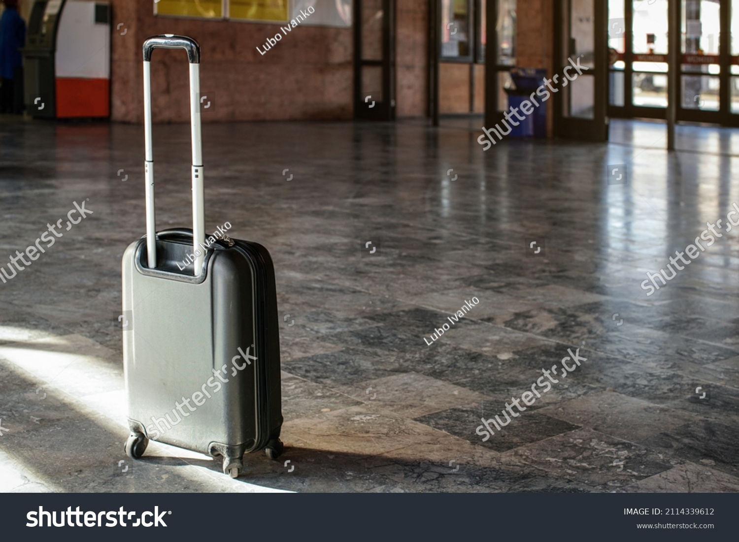 Single unattended trolley wheeled suitcase on stone floor in empty train station hall #2114339612