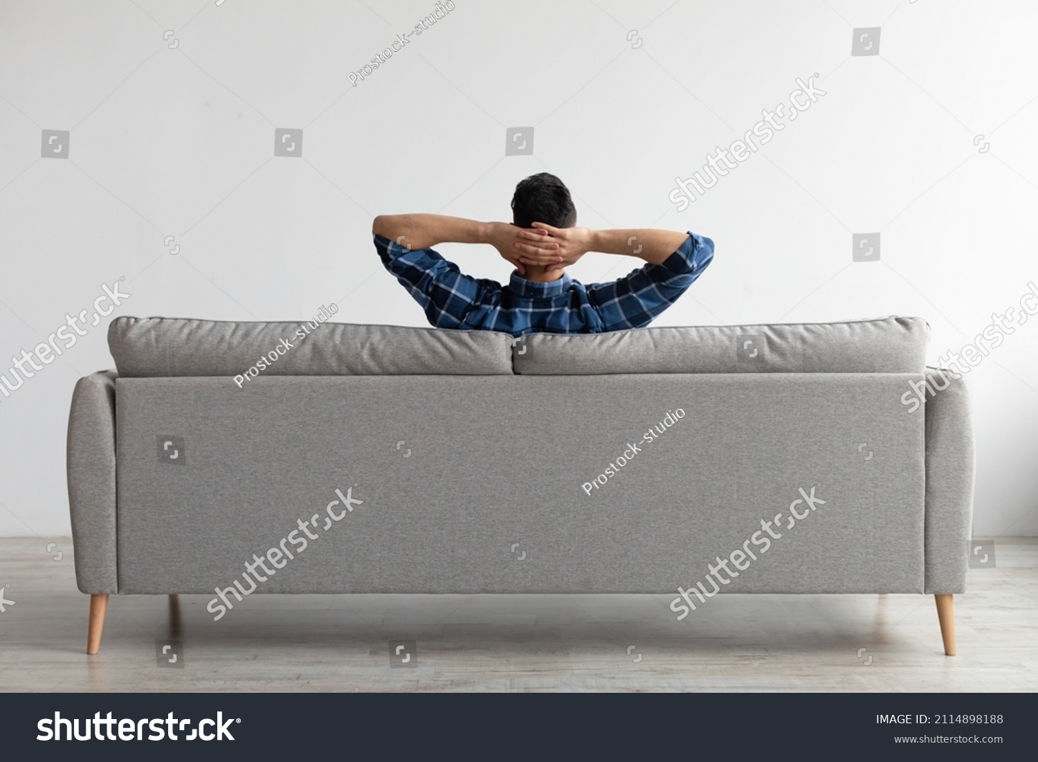Rear view of young guy sitting on comfortable couch at home in living room, looking at wall. Casual man relaxing on sofa, leaning back holding hands behind head, enjoying weekend free time or break #2114898188