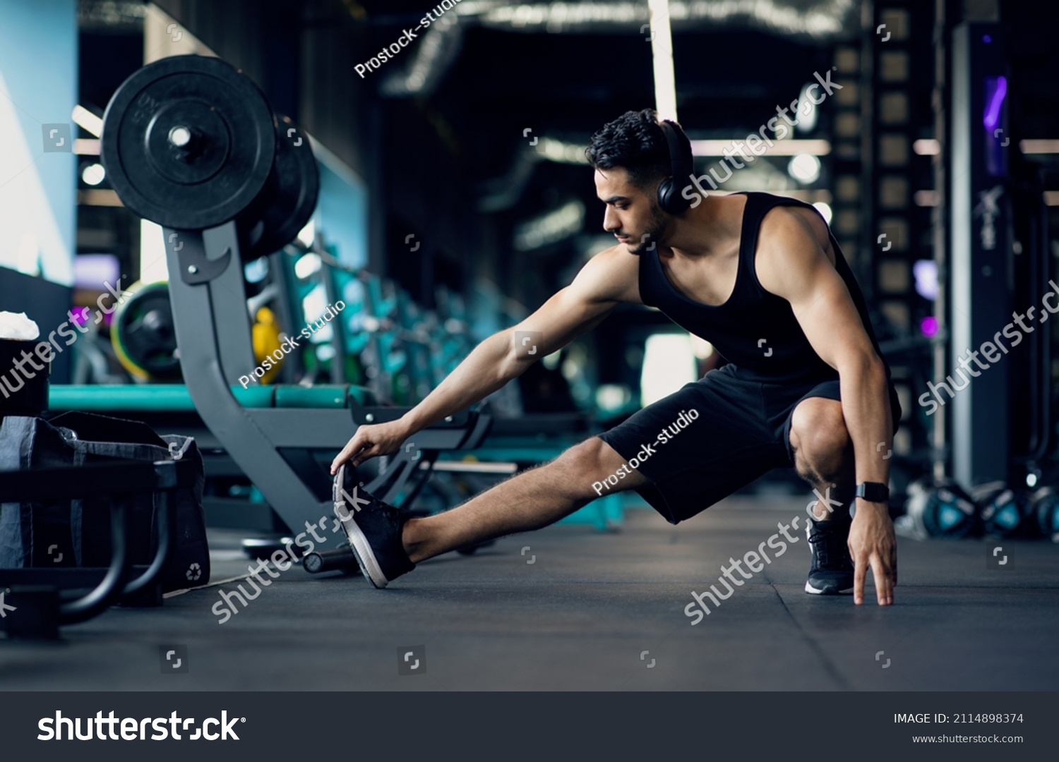 Handsome Young Middle Eastern Male Athlete Warming Up Before Training At Gym, Motivated Millennial Arab Man Stretching Leg Muscles, Preparing For Fitness Workout In modern Sport Club, Copy Space #2114898374
