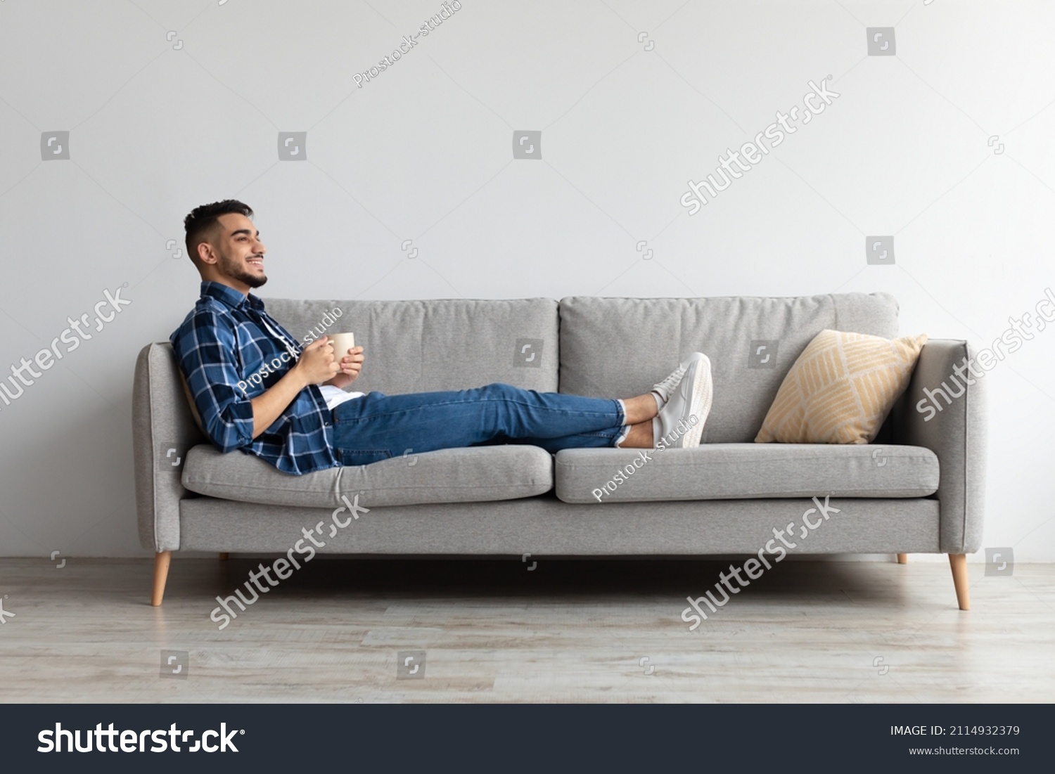Rest Concept. Happy Arab guy drinking coffee sitting on comfortable couch at home in living room. Cheerful casual man relaxing on sofa, enjoying weekend free time or break from work, full body length #2114932379