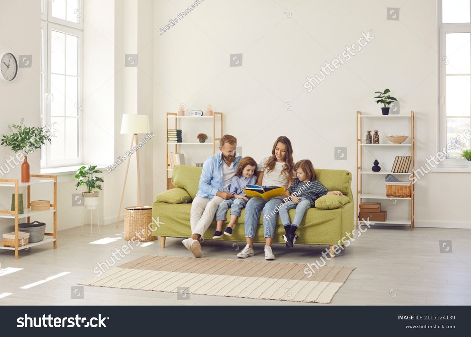 Happy family spending time at home. Mum, dad and little kids reading book while sitting together on green sofa in room with light walls, beige rug and simple brown shelves in their big new house #2115124139