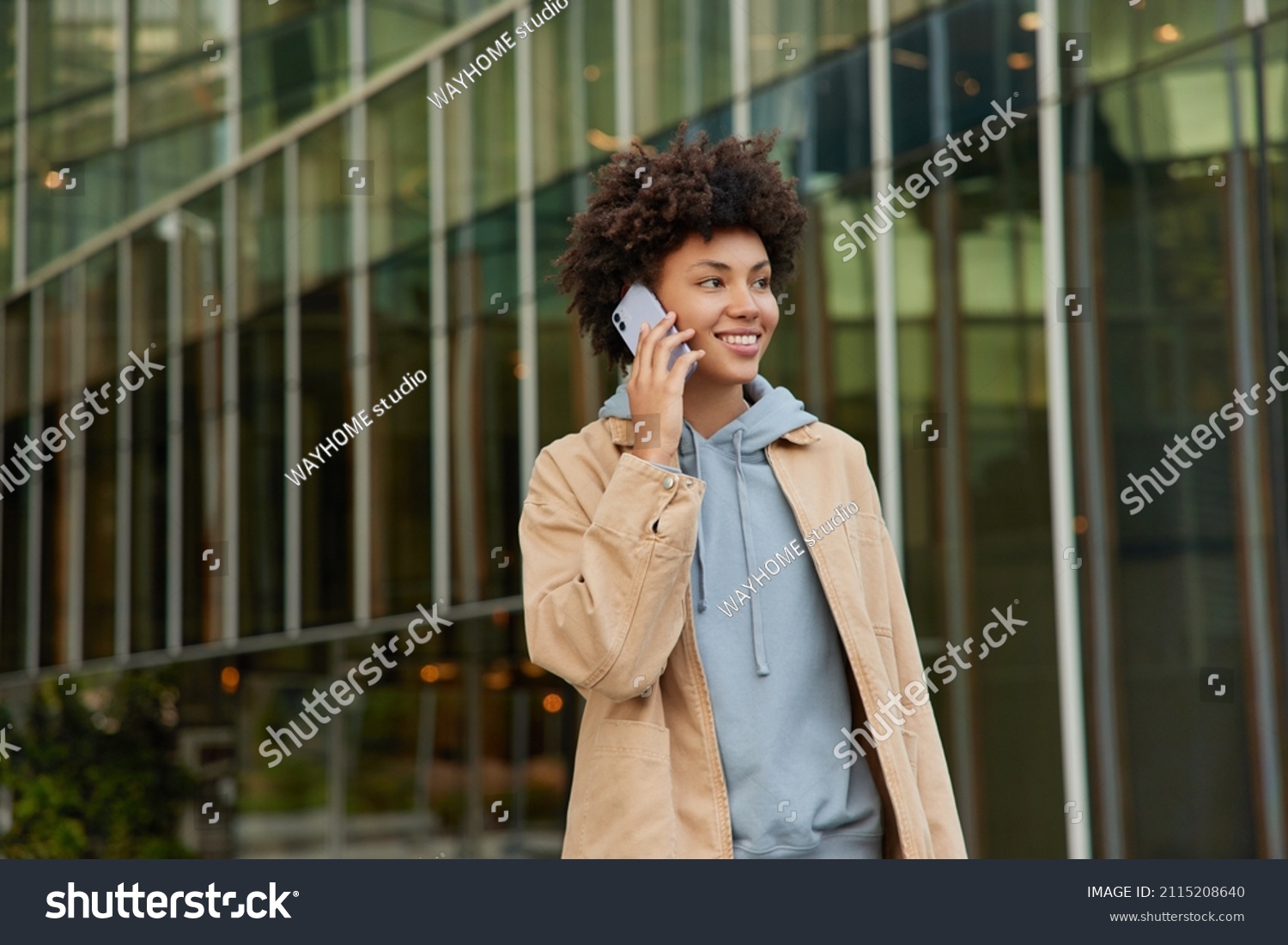 Horizontal shot of happy young woman makes telephone call in roaming wears casual sweatshirt and beige jacket had glad expression walks against blurred city building enjoys smartphone talking #2115208640