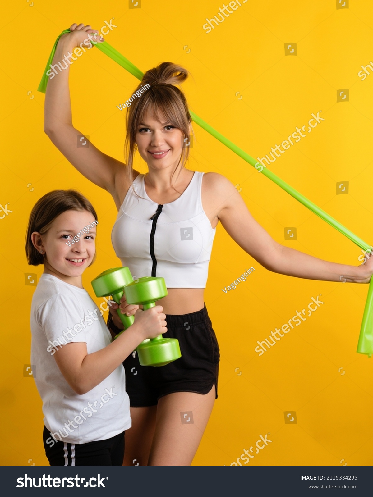 Healthy fit mom and daughter wearing sport wear in studio using resistive exercise band and dumbbells against yellow background. Active lifestyle. Sporty Family together online workouts at home. #2115334295