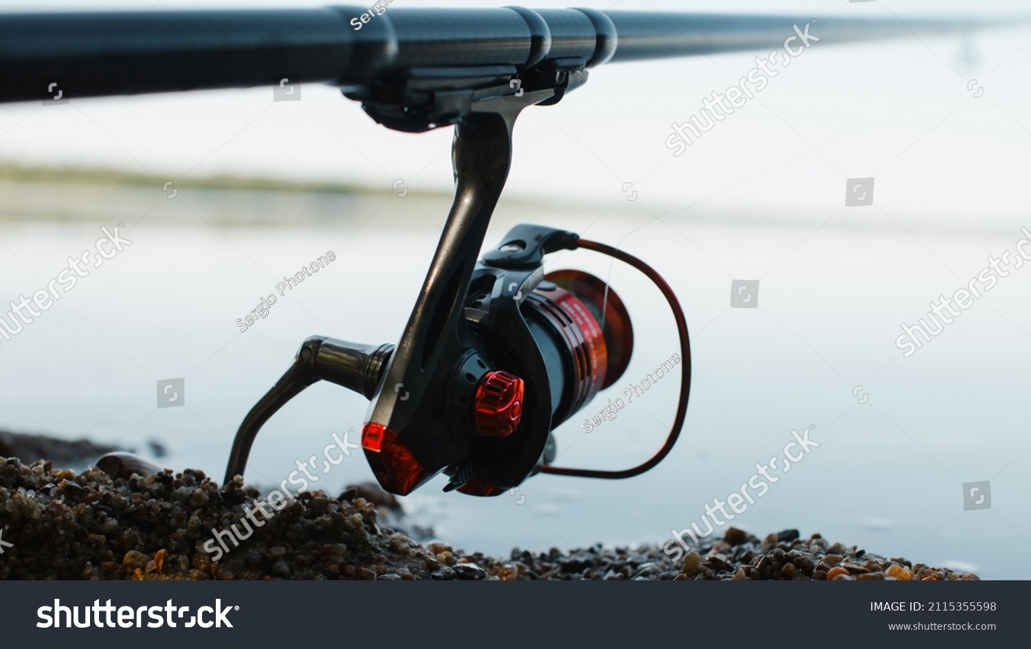 Spinning rod on shore near water outdoors, close-up of steel red reel. Fishing hobby, sport and leisure concept. Selective soft focus. #2115355598