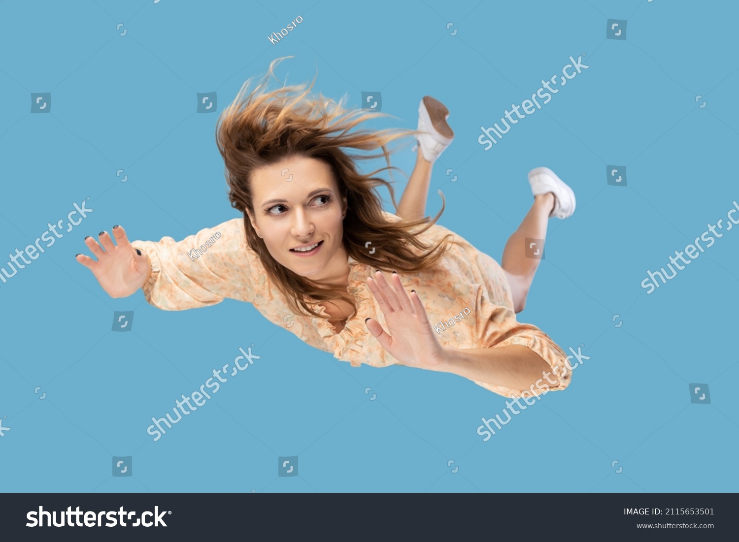 Beautiful young woman levitating in mid-air, falling down and her hair messed up soaring from wind, model flying hovering with dreamy peaceful expression. indoor shot isolated on blue background #2115653501