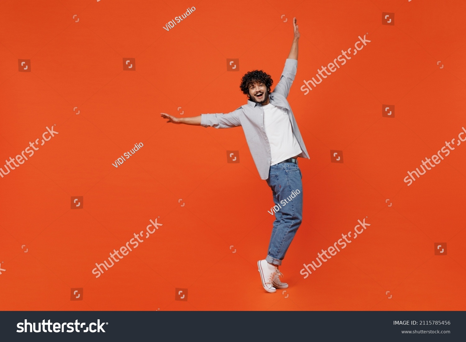 Full size body length smiling young bearded Indian man 20s years old wears blue shirt standing on toes dancing lean back have fun spreading hands isolated on plain orange background studio portrait #2115785456