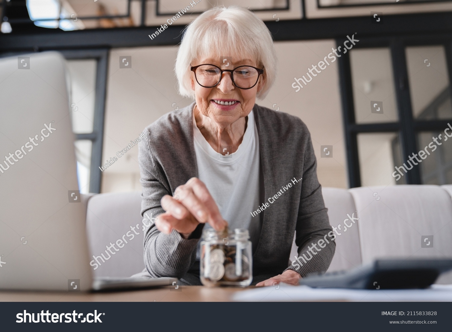 Savings concept. Nest egg of old elderly senior woman grandmother saving money, economizing pension, mortgage loan at home using laptop and putting coin into moneybox #2115833828
