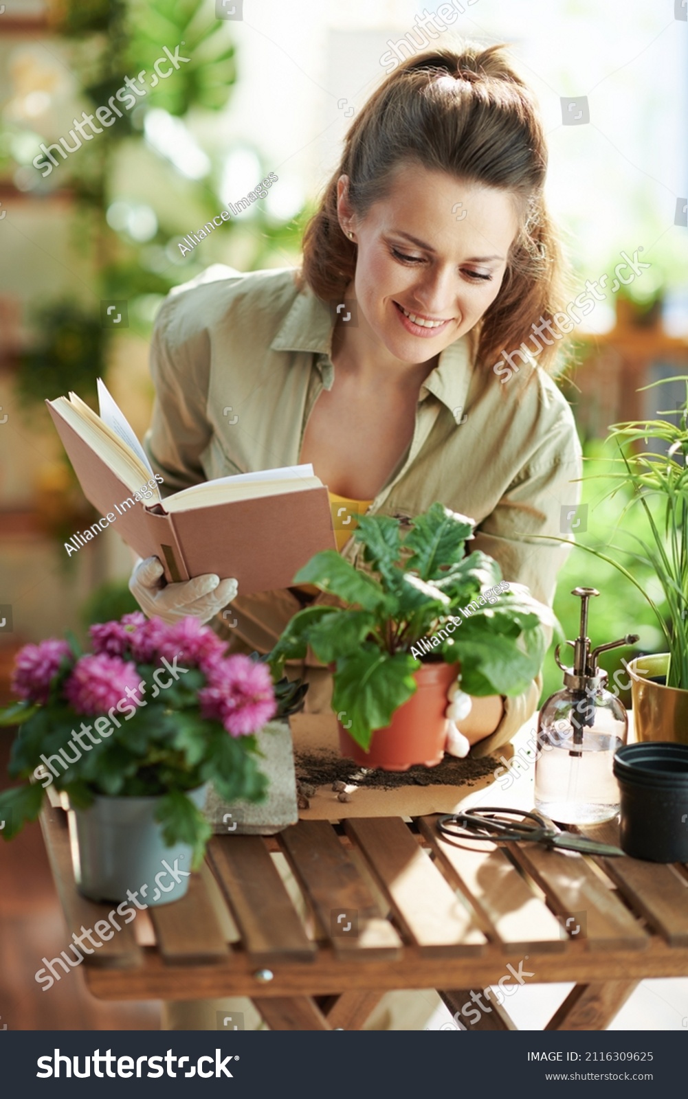 Relaxing home gardening. happy modern middle aged housewife in white rubber gloves with potted plant and book at modern home in sunny day. #2116309625
