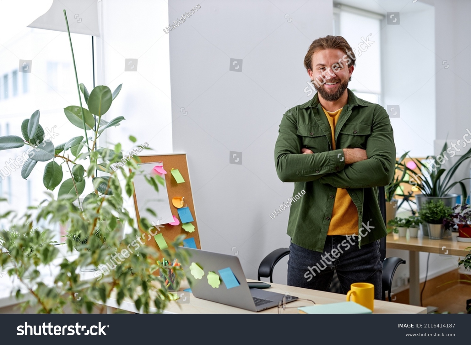 Portrait of excited caucasian manager designer man in office posing at camera, standing behind desk with laptop, many plants in office room. European guy in casual wear, success and business #2116414187