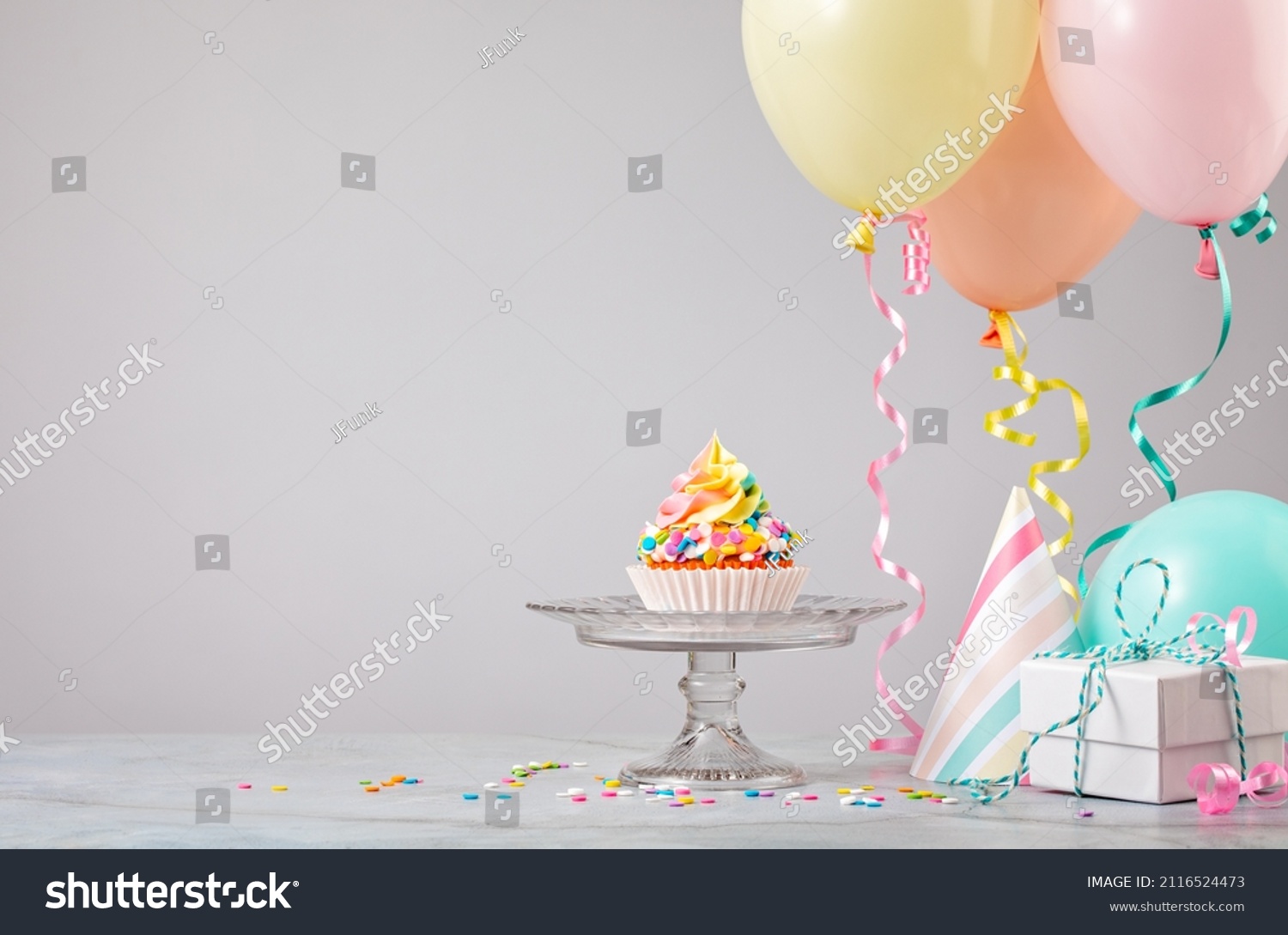 Rainbow Birthday cupcake on a stand with presents, hats and colorful balloons over light grey background. Scene from a birthday party! #2116524473