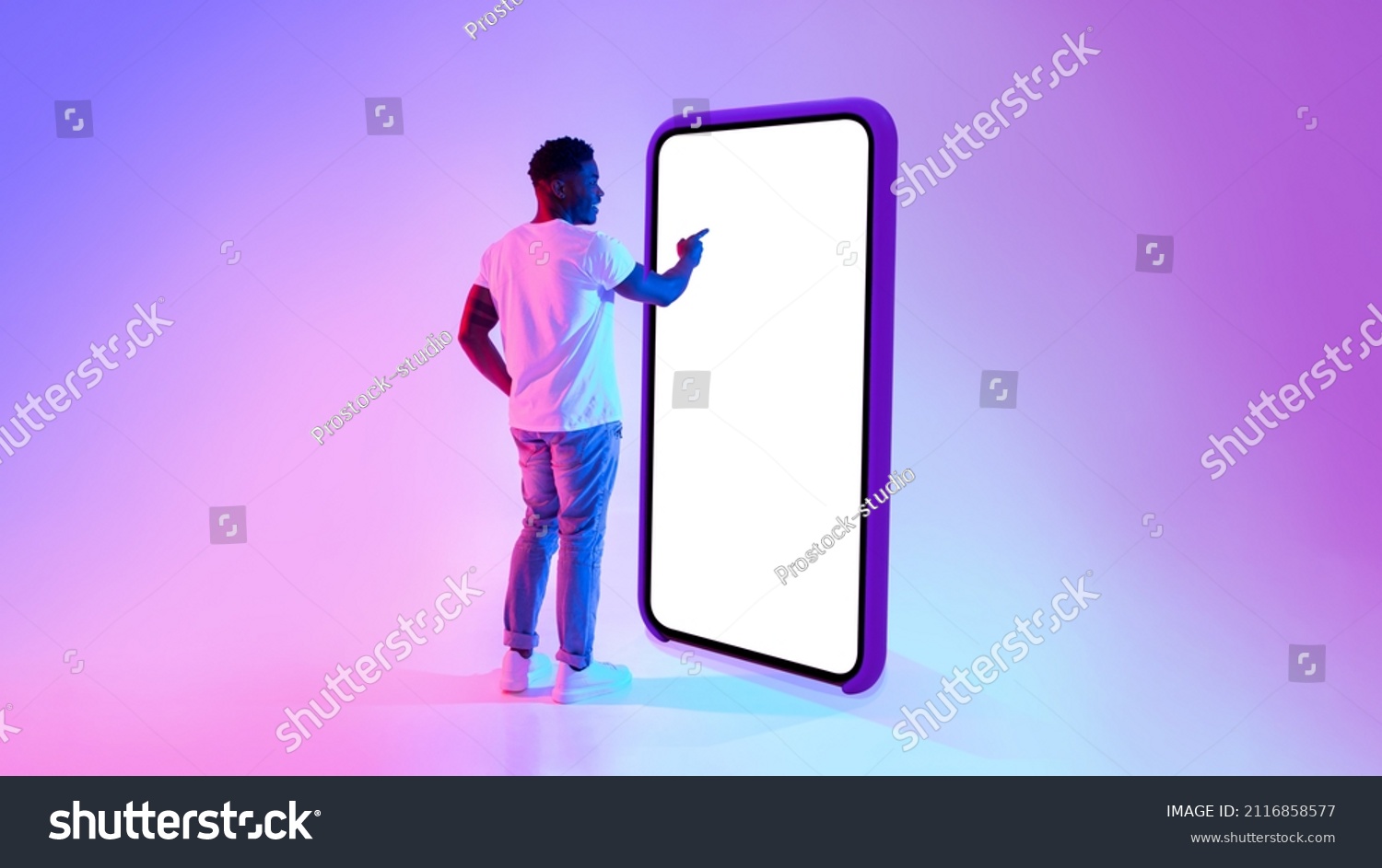 Full length of millennial black guy standing near big cellphone with mockup for your app on screen, interacting with user interface in neon light. Smartphone display template for website or ad #2116858577