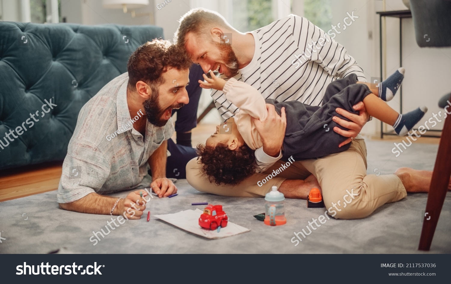 Loving LGBTQ Family Playing with Toys with Adorable Baby Boy at Home on Living Room Floor. Cheerful Gay Couple Nurturing a Child. Concept of Diverse Childhood, New Life, Parenthood. #2117537036