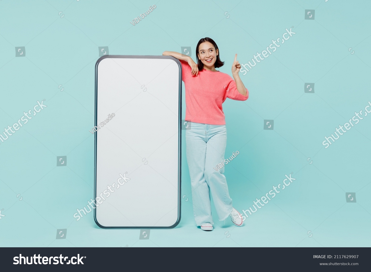 Full body young woman of Asian ethnicity in pink sweater stand near big mobile cell phone with blank screen workspace area point finger up with new idea isolated on pastel plain light blue background #2117629907