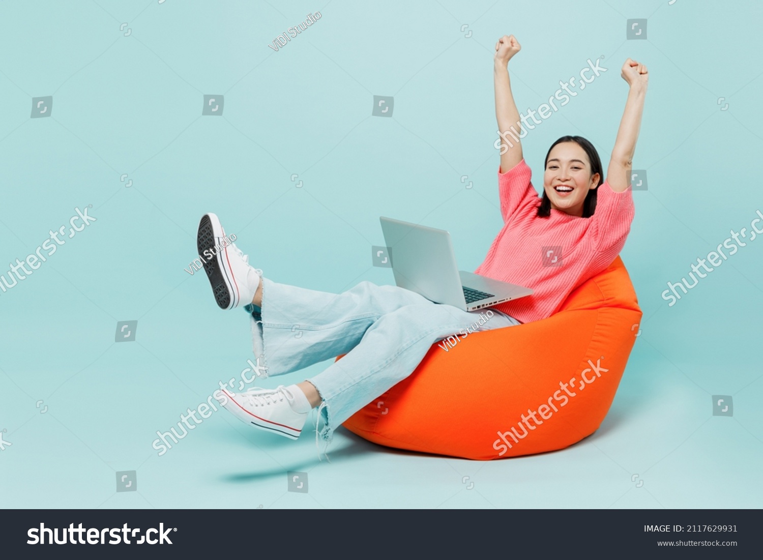 Full body young happy woman of Asian ethnicity 20s in pink sweater sit in bag chair use work on laptop pc computer with outstretched hands finish job isolated on pastel plain light blue background. #2117629931