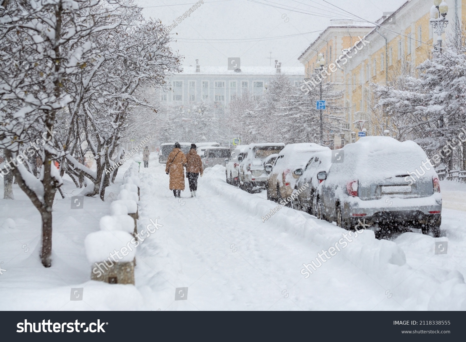 Snow covered city street during a heavy snowfall. Lots of snow on the sidewalk, cars and tree branches. Women walk around the winter city. Cold snowy weather. Magadan, Siberia, Russian Far East. #2118338555