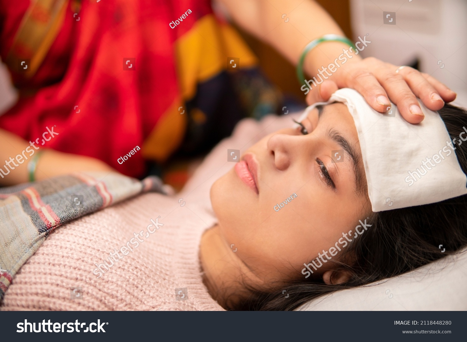 Indoor image of Indian matures mother sponging her ill daughter’s forehead to reduce the temperature down. She is having a high fever and lying in bed at home. She is wearing warm clothes. #2118448280