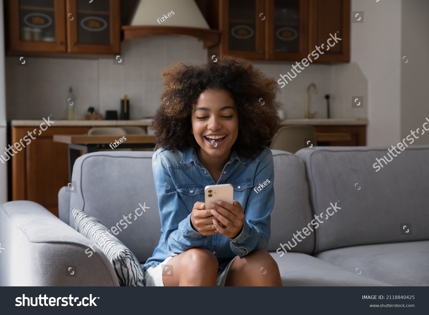 Cheerful happy gen Z teen girl receiving exciting message on smartphone, reading text in screen, smiling, laughing, sitting on couch, using online virtual app on mobile phone #2118840425