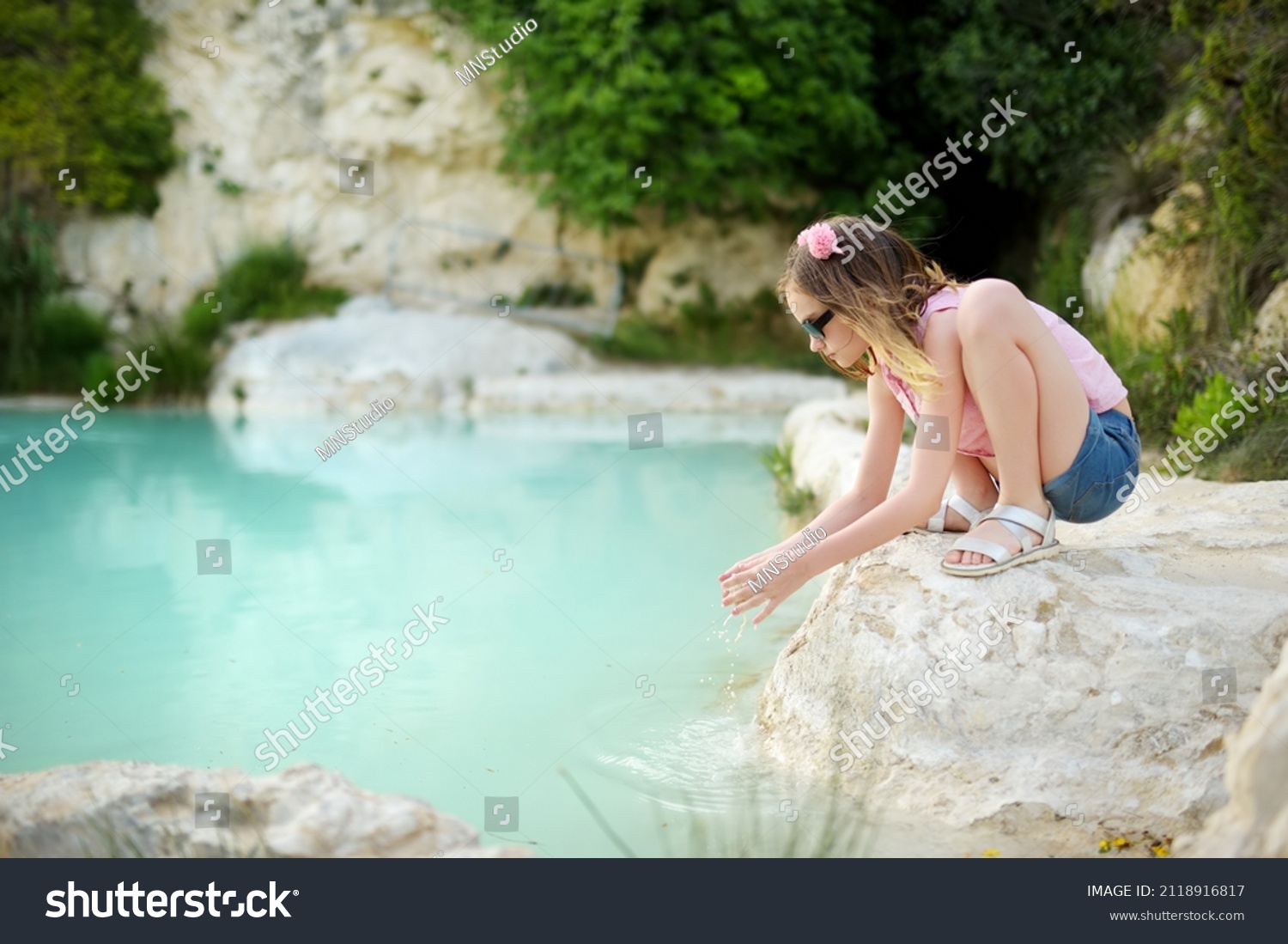 Young girl playing by natural swimming pool in Bagno Vignoni, with thermal spring water and calcium carbonate deposits, which form white concretions and waterfall. Tuscany, Italy. #2118916817