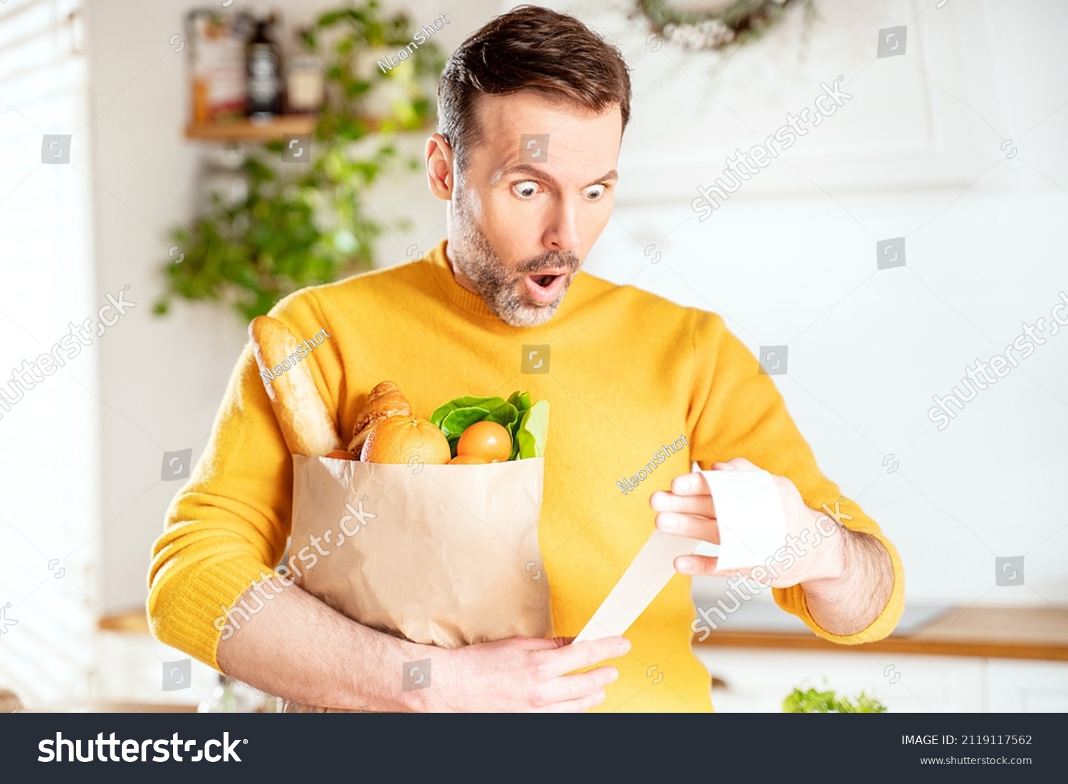Surprised man looking at store receipt after shopping, holding a paper bag with healthy food. Guy in the kitchen. Real people expression. Inflation concept. #2119117562